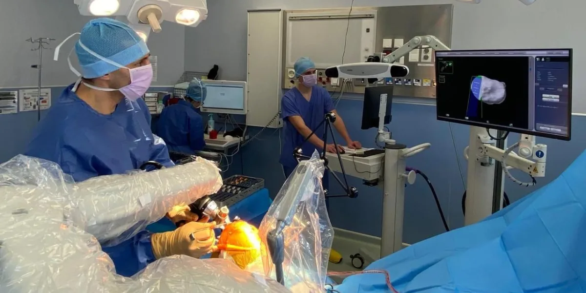Personne en tenue bleue et masque, mains gantées sur un bras articulé sous housse plastique, écran 3D à droite