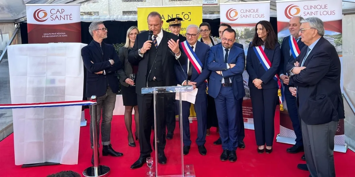 Homme en costume parlant au micro derrière un pupitre transparent, entouré de personnes en écharpe tricolore